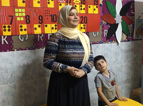 A teacher stands in front of the class with a young student by her side.